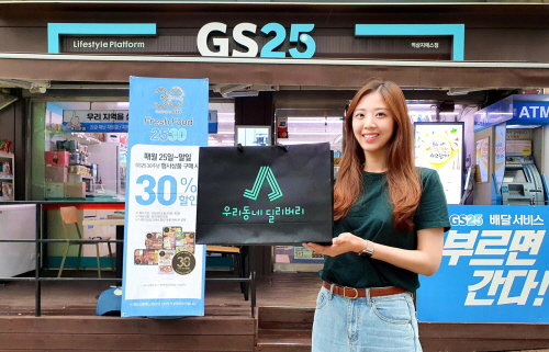 모델이 GS25 앞에서 우리동네딜리버리 배송 홍보물을 들고 포즈를 취하고 있다